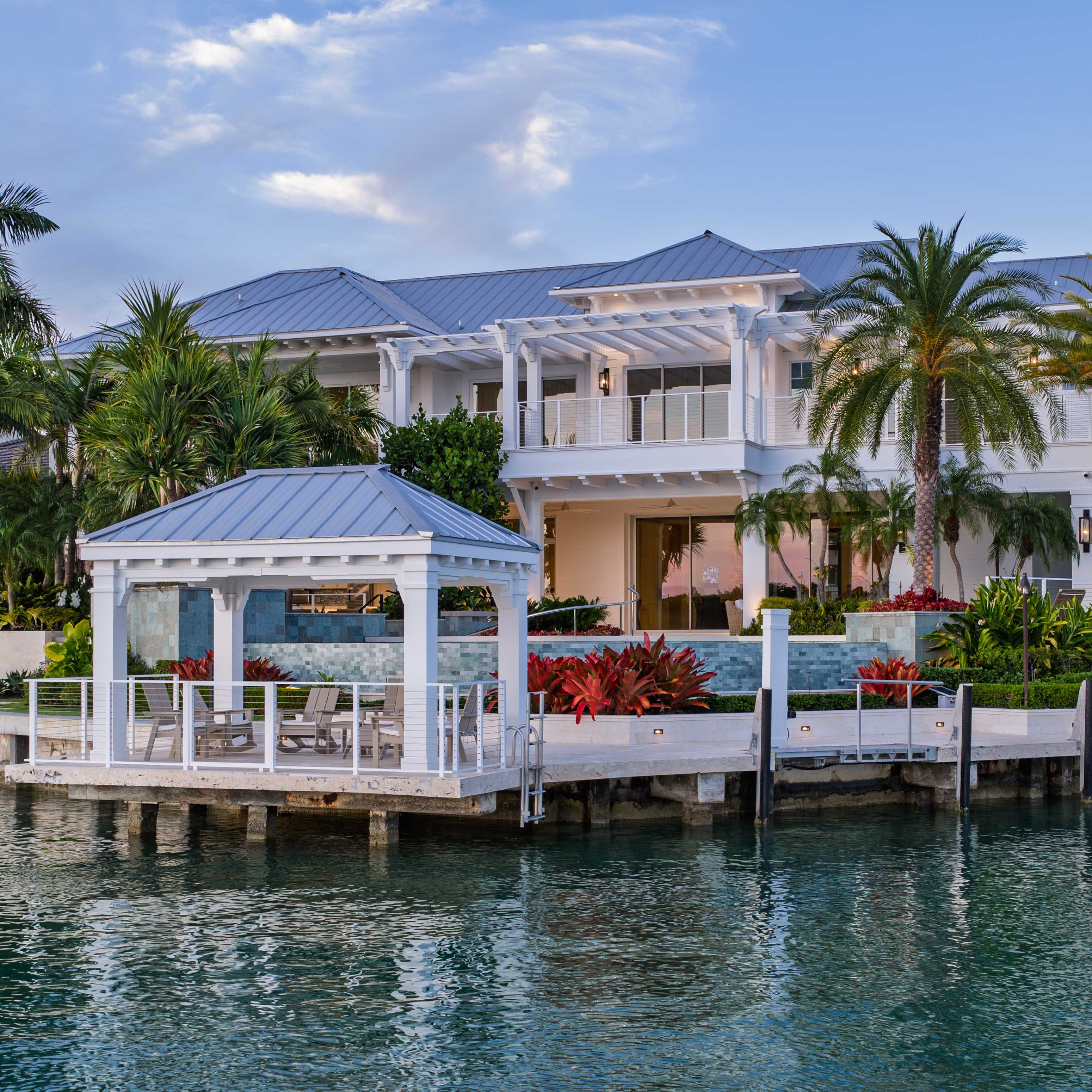 A waterfront home with white terraces, tropical planting and a dock at dusk.