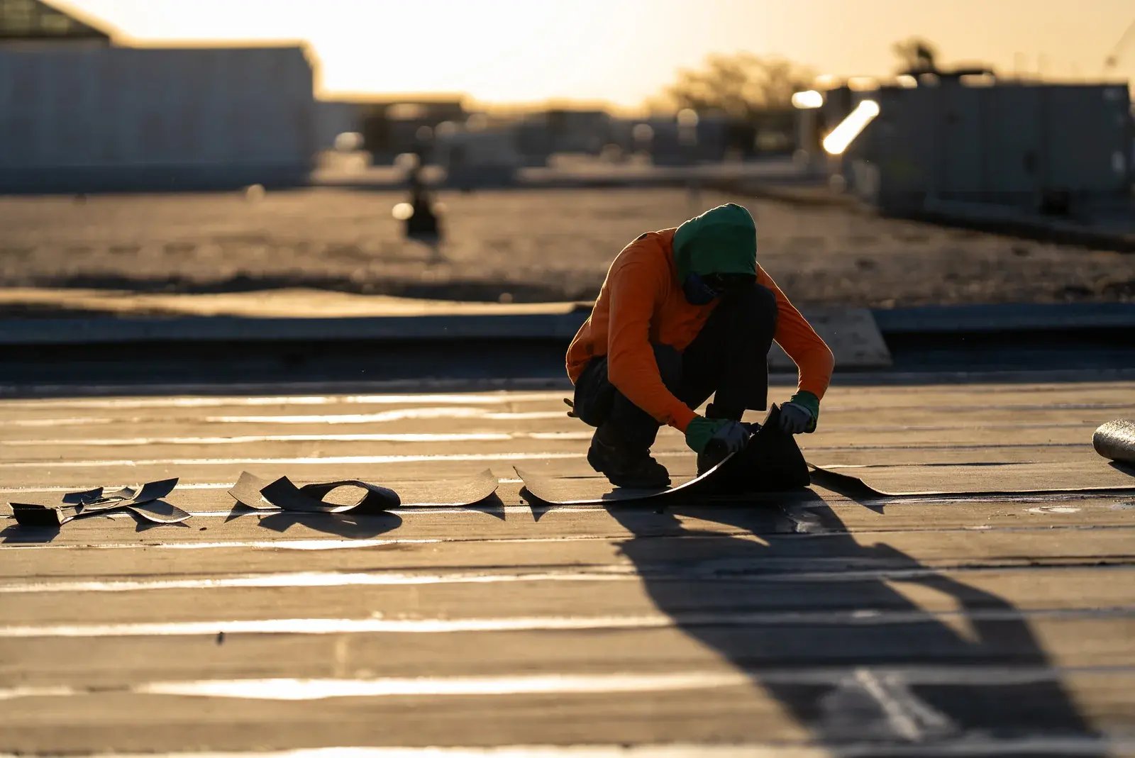 A roofer kneeling to work on a roof membrane in warm low sunlight.