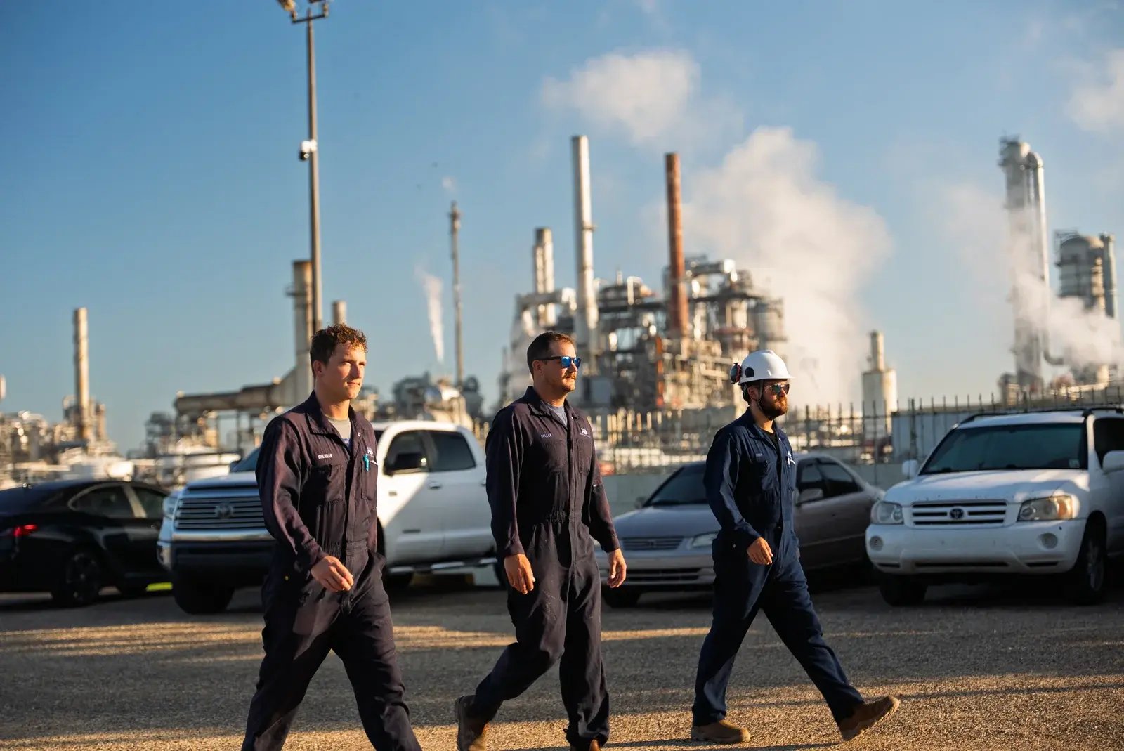 Three people walking through an industrial facility beside parked trucks.