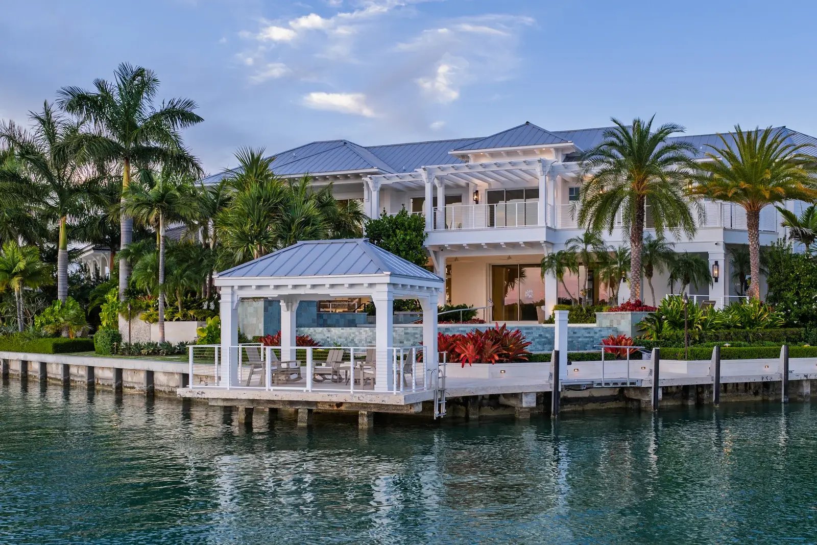 A waterfront home with white terraces, tropical landscaping and a dock at dusk.