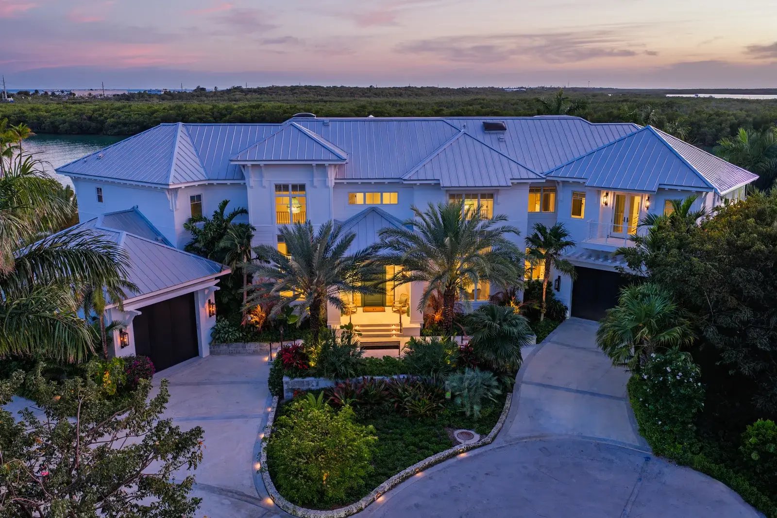 The illuminated entrance and palm-lined drive of a coastal home at dusk.