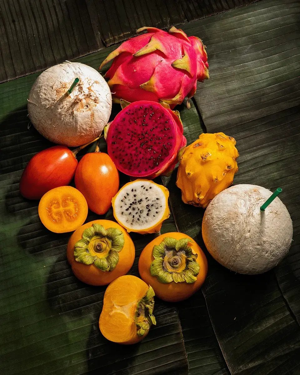 Dragon fruit, coconuts and cut tropical fruit arranged on dark green leaves.